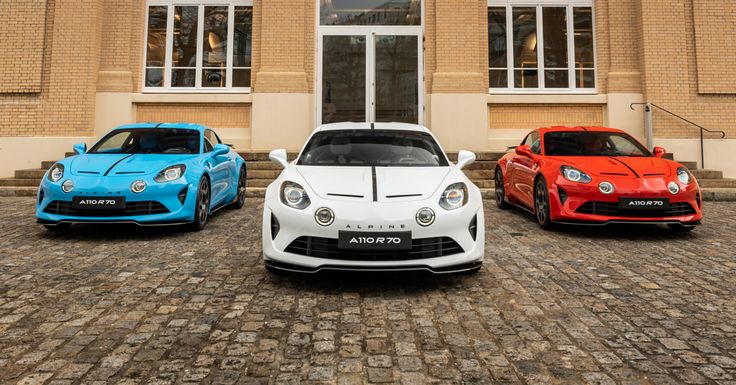 Three Alpine A110 sports cars in blue, white, and red parked on cobblestone in front of a building.