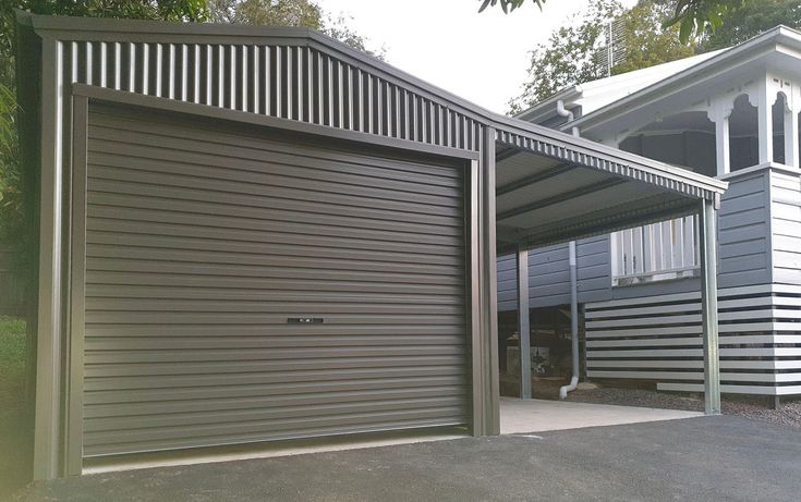 Steel garage with a roller door and carport next to a blue house, set among trees.