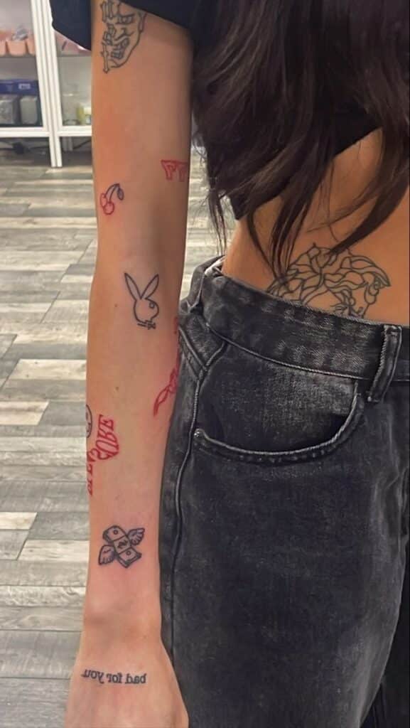 Arm and waist tattoos featuring cherries, money, and designs on a person in black jeans and top on a wood floor.