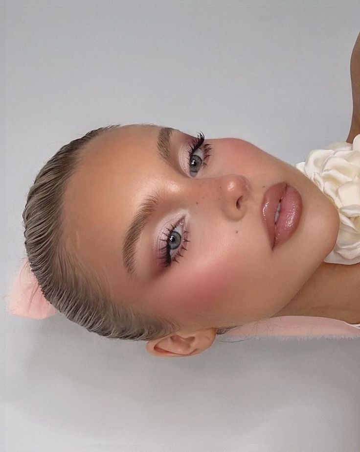 Close-up of a model with dewy skin, pink blush, glossy lips, and sleek hairstyle, showcasing elegant makeup.