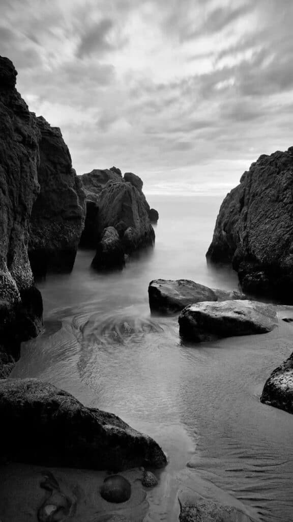 Monochrome coastal scene with large rocks, gentle waves, and a cloudy sky, creating a serene, natural landscape view.