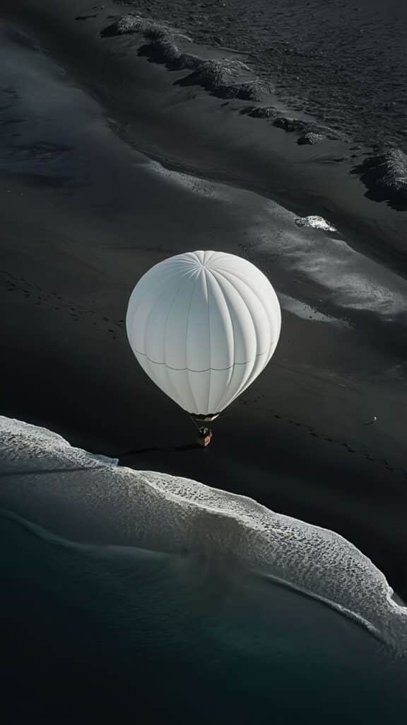 White hot air balloon floating over a black sand beach near ocean waves.