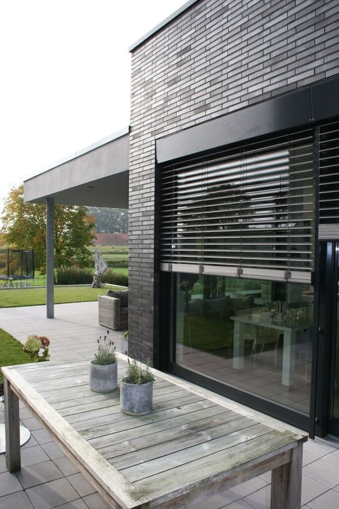 Modern patio with wooden table, potted plants, and large window blinds against a brick wall.