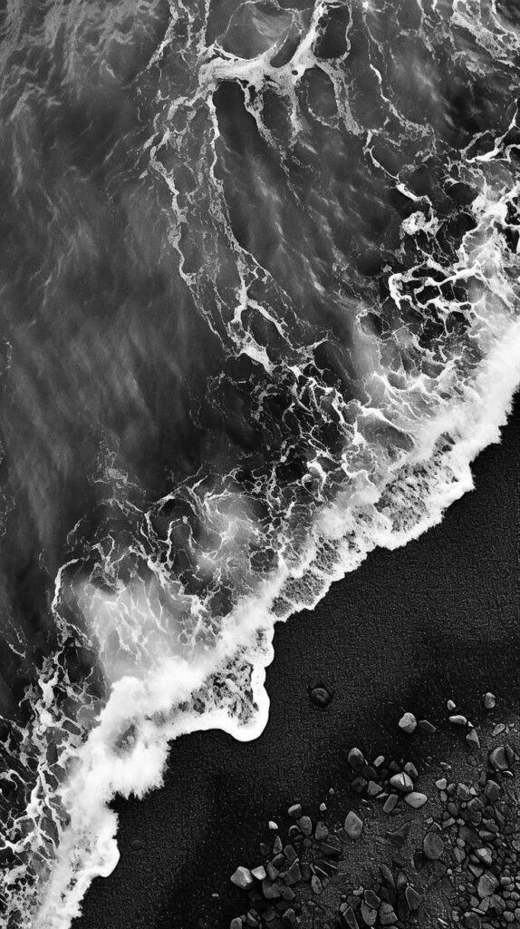 Black and white aerial view of ocean waves crashing on a rocky beach.