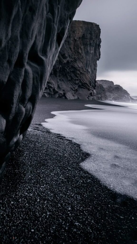 Dramatic coastal scene with black sand beach, towering cliffs, and foamy waves under a moody gray sky.