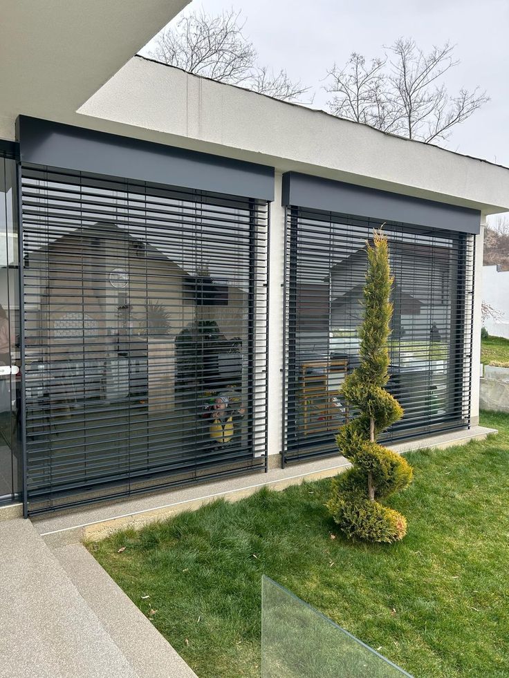 Modern building facade with large glass windows, horizontal blinds, and a neatly trimmed spiral shrub in the green lawn.