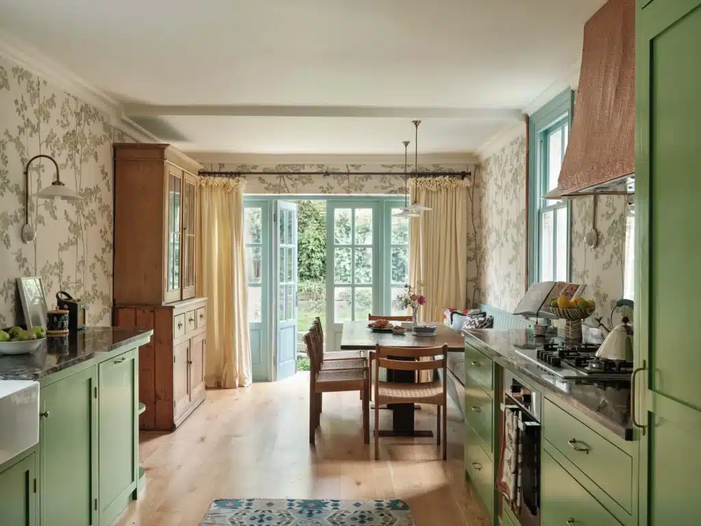 Charming farmhouse kitchen with green cabinets, wooden dining set, and floral wallpaper leading to garden door.