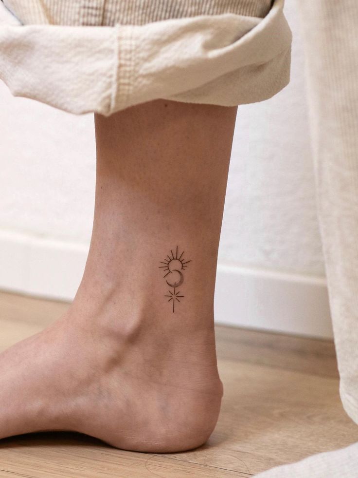 Minimal sun and moon ankle tattoo on person’s leg. Simple, elegant design with celestial theme on wooden floor background.