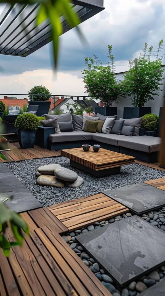 Modern rooftop patio with gray sectional sofa, wooden coffee table, and lush greenery under cloudy sky.