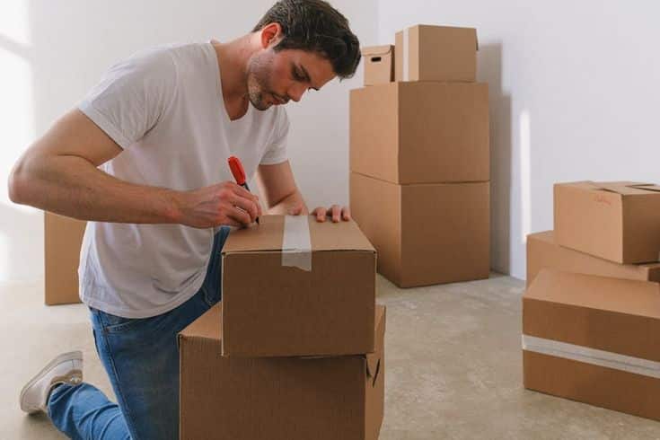 Man labeling cardboard moving boxes in room filled with packed boxes.