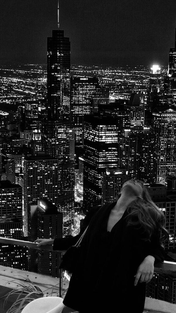Person enjoying a nighttime rooftop view of a city skyline, featuring bright lights and skyscrapers.