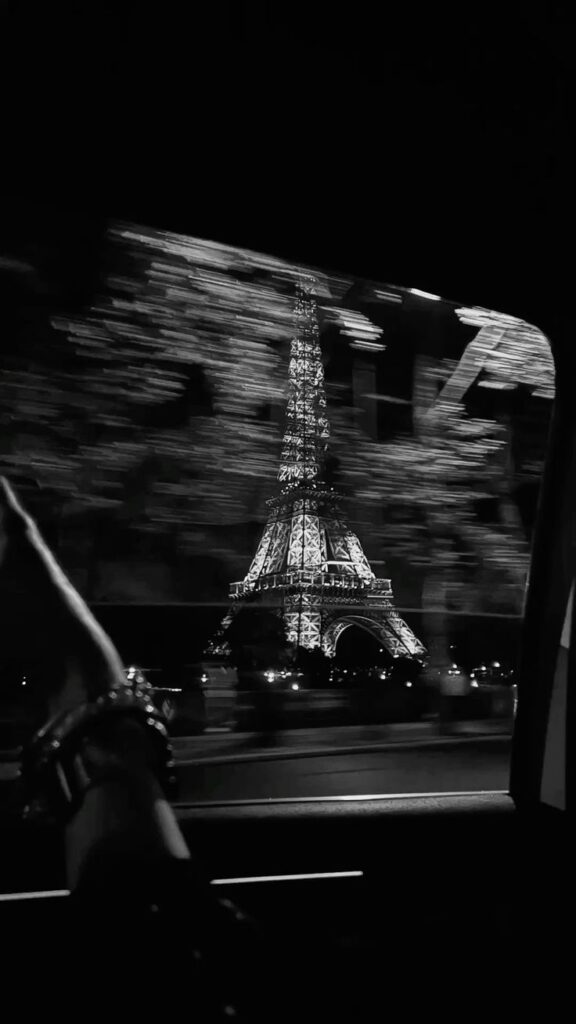 View of illuminated Eiffel Tower at night from a moving car window, with blurred trees in the foreground.