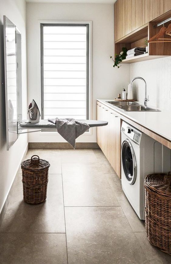 Modern laundry room with washer, ironing board, wicker baskets, and a sink under natural light. Clean and organized.