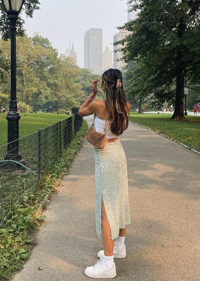 2af343ec-9c62-473b-93fb-9b4cd6ddfbef Woman in white top and floral skirt walking in a park, surrounded by green trees, with city skyline in the background. | Sky Rye Design Woman in white top and floral skirt walking in a park, surrounded by green trees, with city skyline in the background.