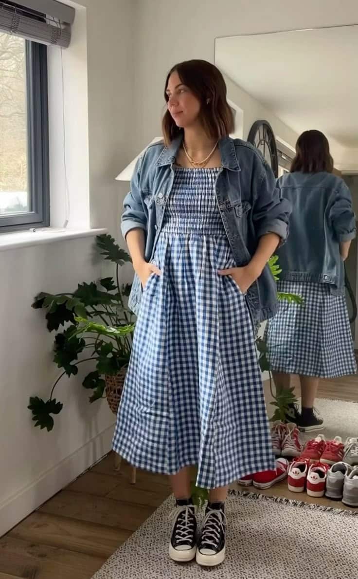 Woman in denim jacket and blue gingham dress stands near window, looking outside, with sneakers and plants nearby.