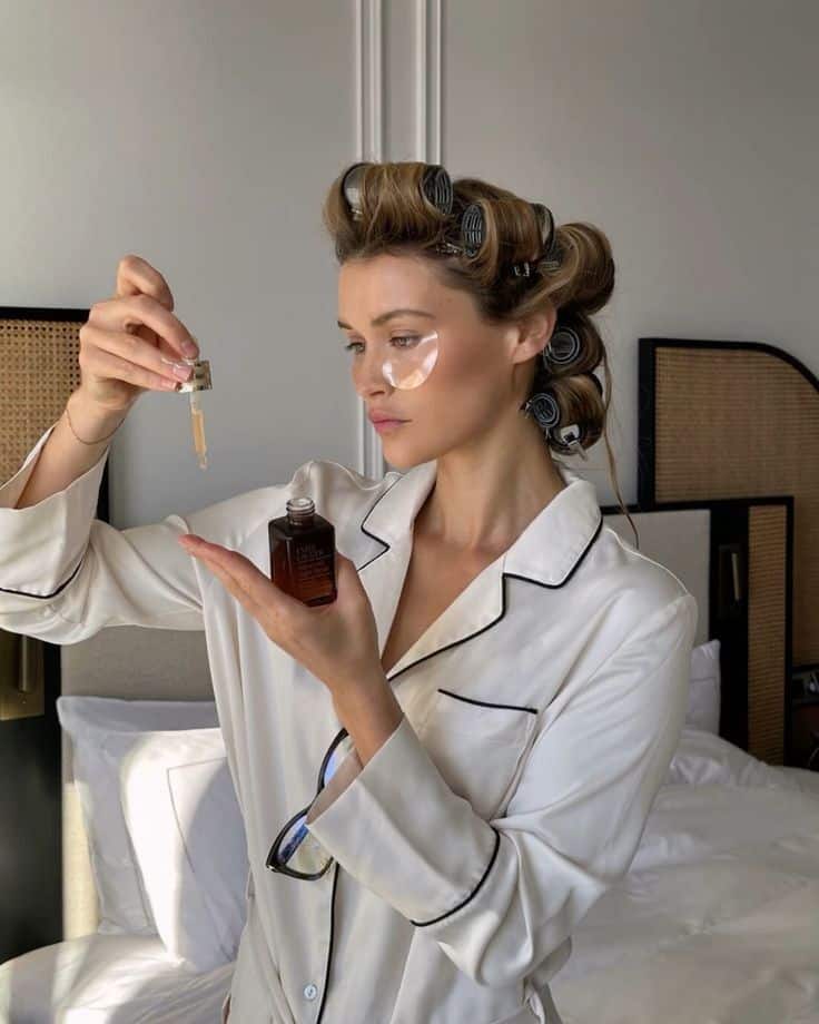 Woman in pajamas applying facial serum with eye patches and hair rollers, enhancing morning skincare routine.