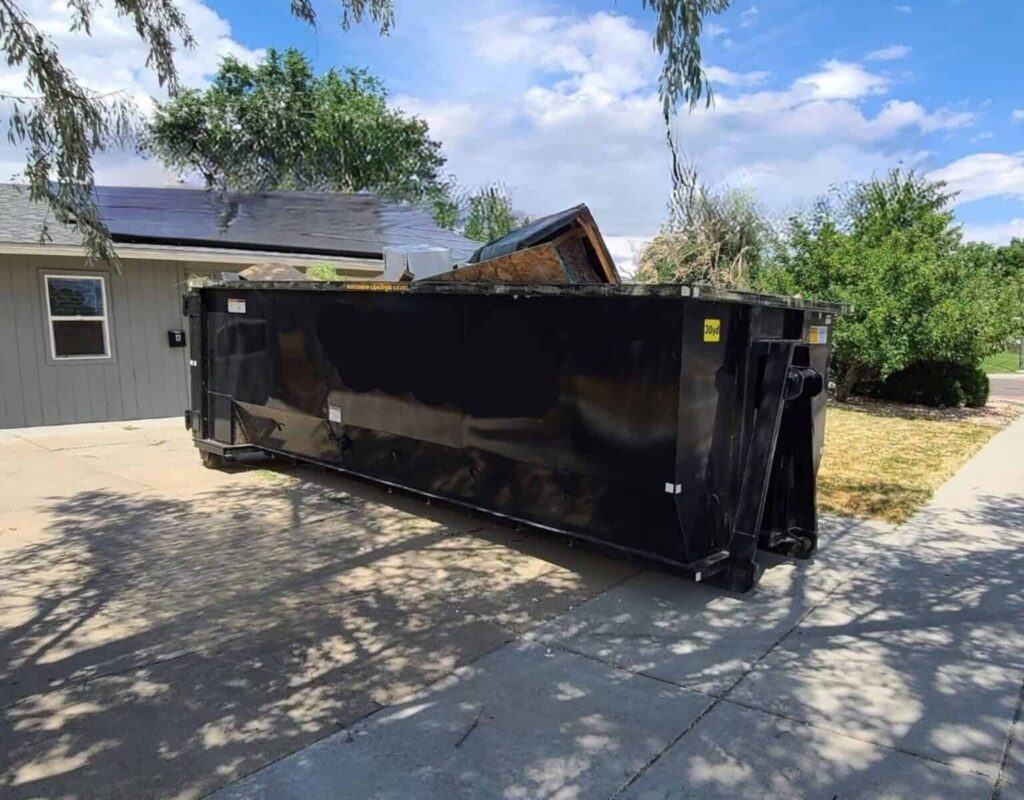 Black dumpster outside house on driveway near trees and bushes under a clear blue sky.