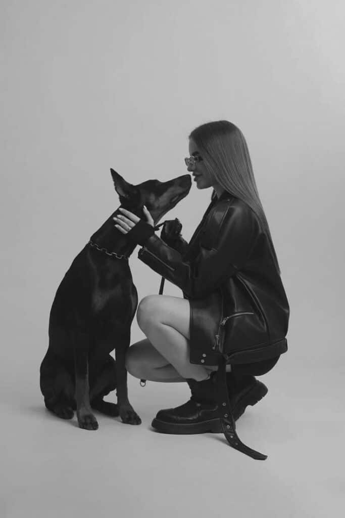Woman in leather jacket kneeling and bonding with black dog in studio, grayscale image.