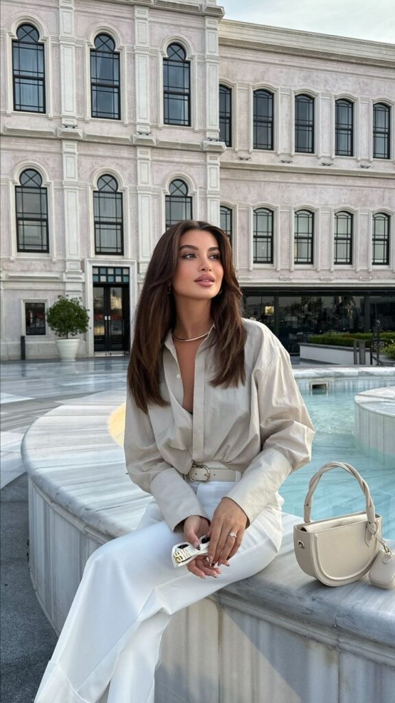 Elegant woman in stylish beige outfit sitting by fountain with classic building background.