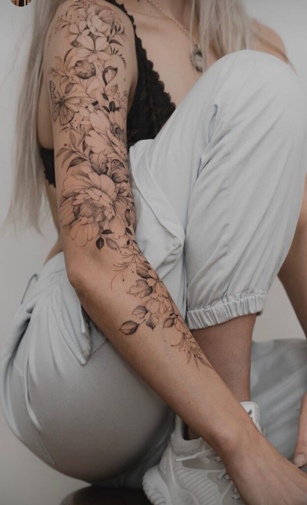 Floral tattoo sleeve on woman's arm with white clothing and sneakers.