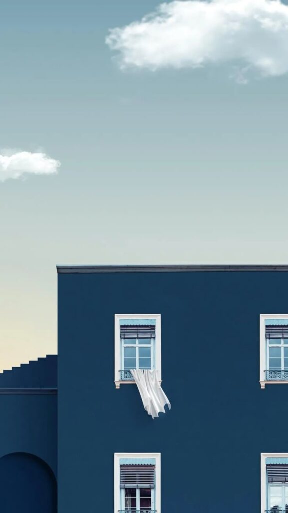 Blue building with open windows and billowing white curtain under a clear sky with clouds.