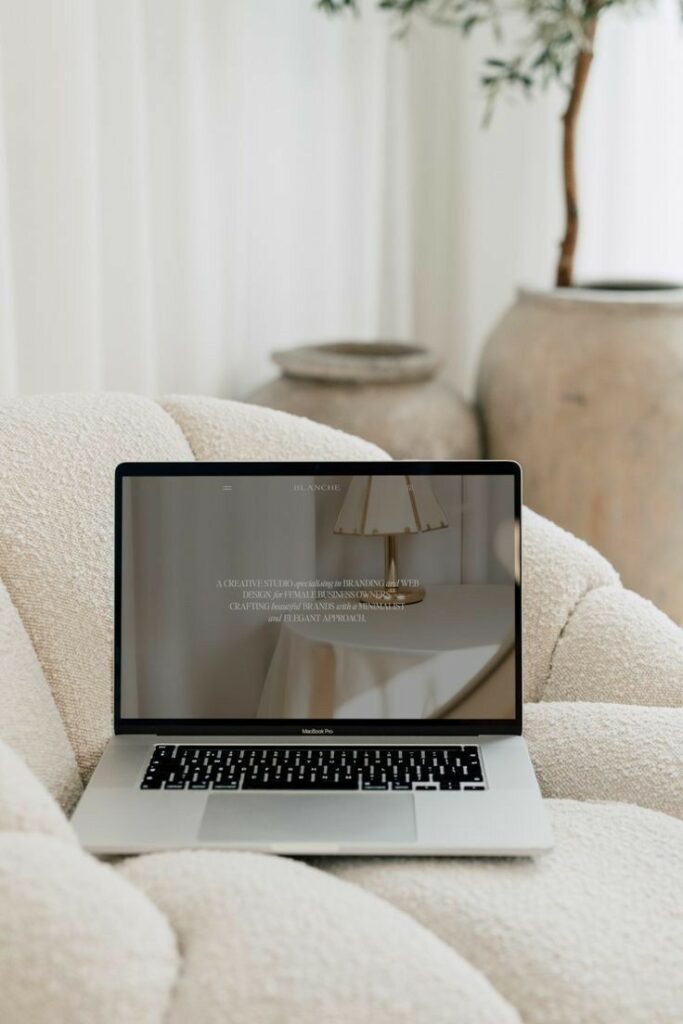 Laptop on a cozy chair displaying minimalist interior design website, with pottery and plants in the background.