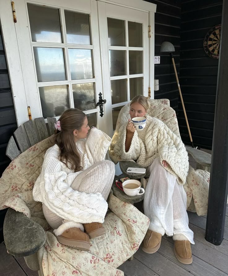 Two people in cozy sweaters and slippers enjoying hot drinks on a porch with floral blankets.