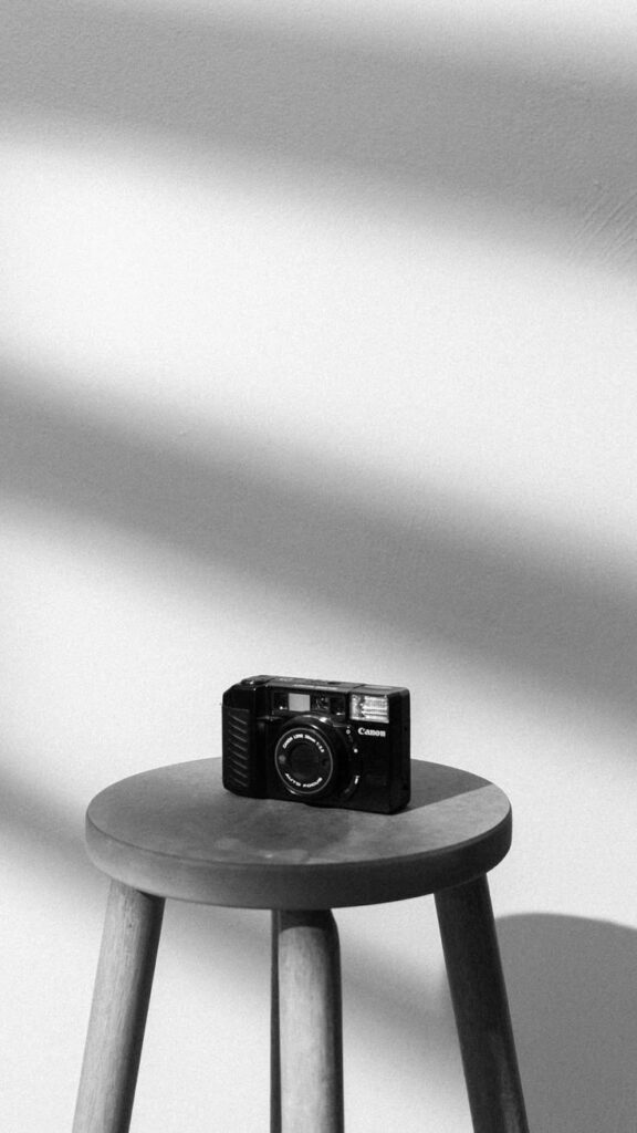 Black vintage camera on a wooden stool with shadowed wall background in black and white.