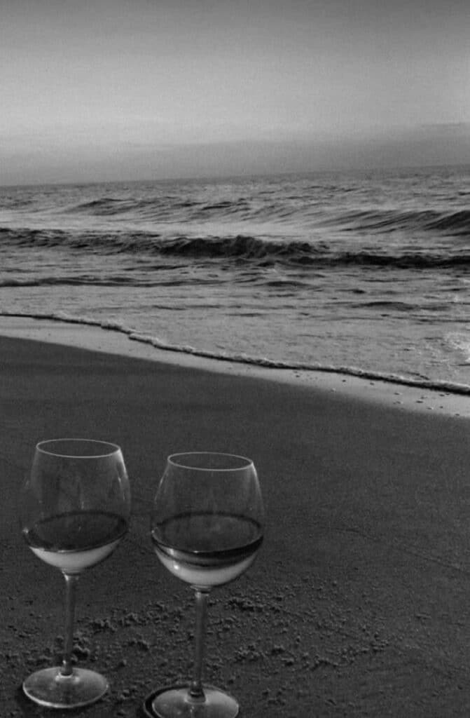 Two wine glasses on sandy beach with ocean waves in background, creating a romantic seaside atmosphere.