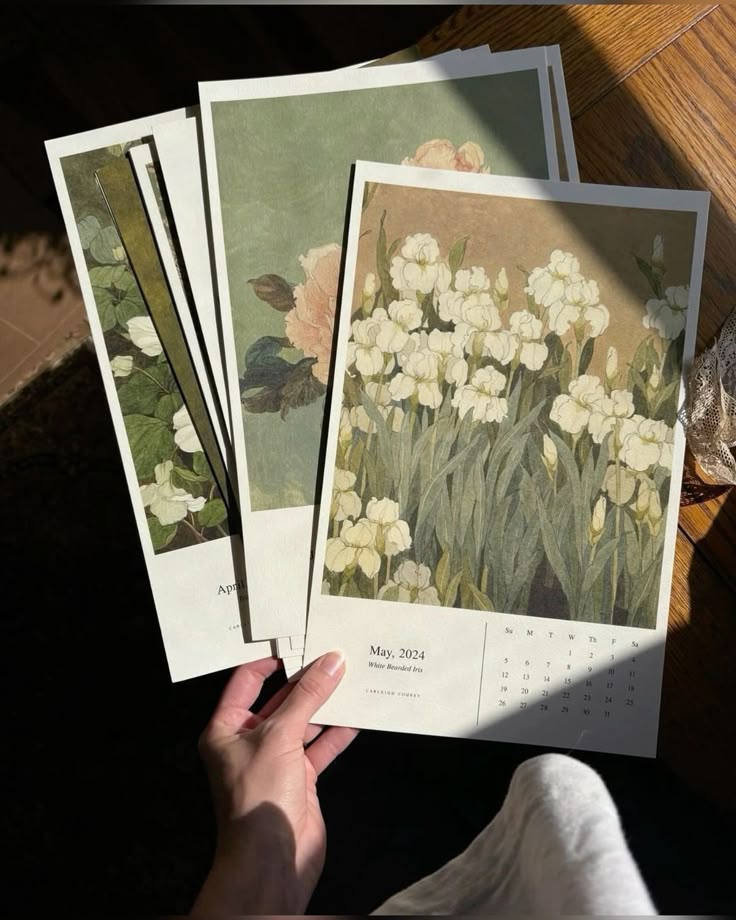 Hand holding floral 2024 calendars with botanical prints in sunlight on a wooden table.