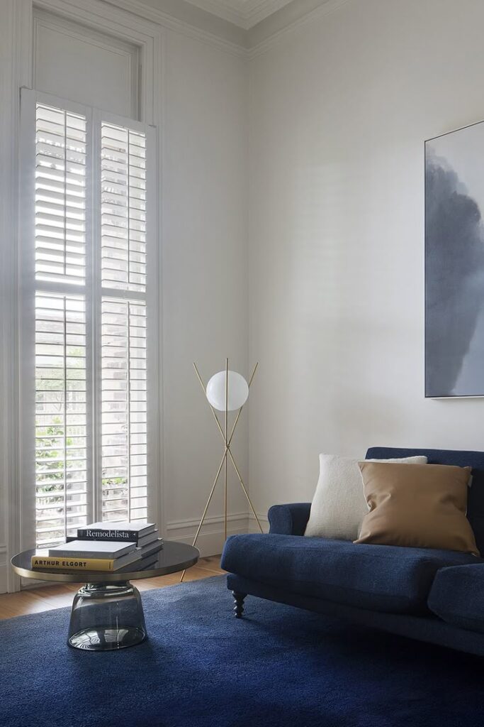 Modern living room with blue sofa, white walls, and stylish floor lamp by a large window with shutters.