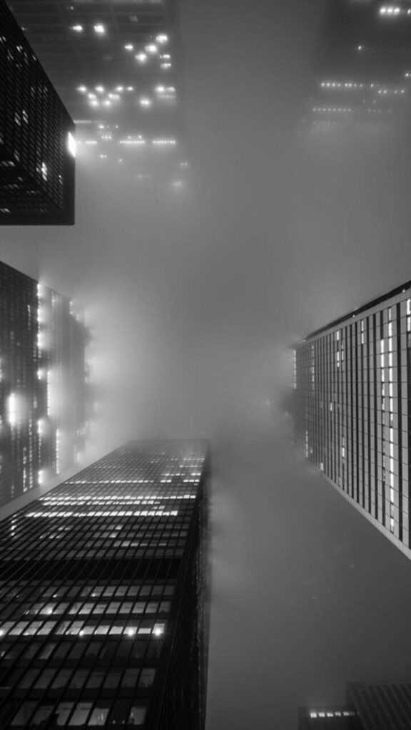 Foggy city skyline with tall skyscrapers disappearing into the misty night, illuminated by glowing windows.
