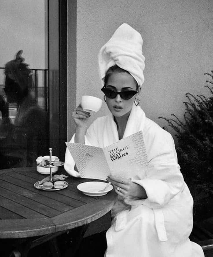 Woman in robe with towel turban reading a card, sipping coffee at outdoor table with macarons, in a chic black and white style.