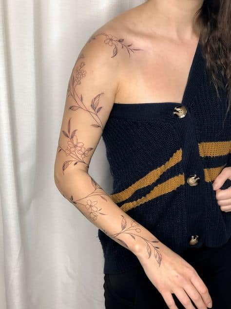 Woman with floral arm tattoos wearing a navy sweater with yellow stripes, posing against a white curtain.