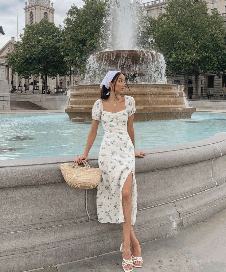 Woman in floral dress by a fountain, holding straw bag and wearing white sandals, outdoor city setting.