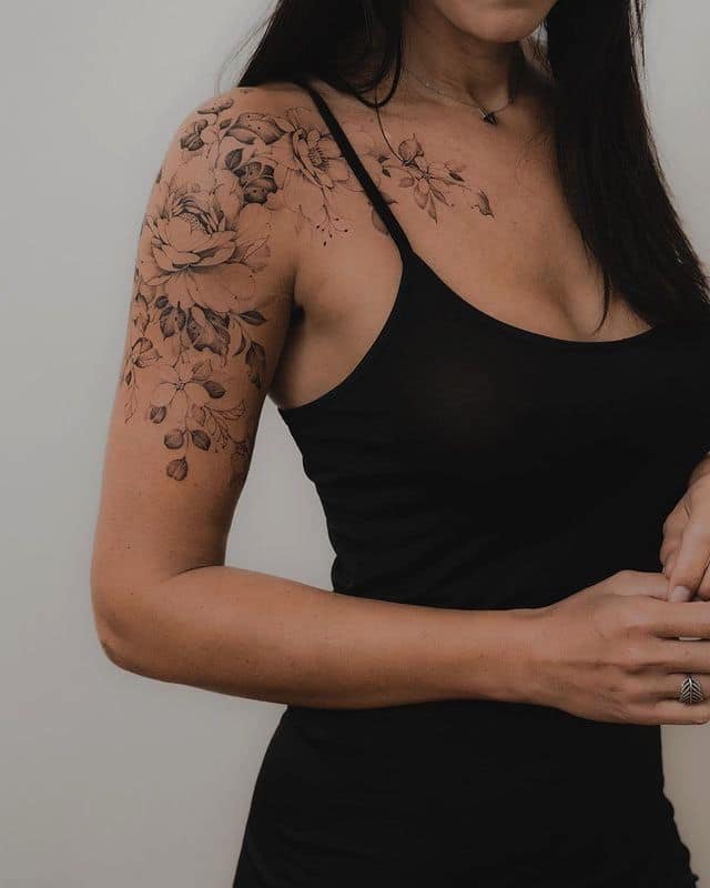 Woman in black dress with floral shoulder tattoo, standing against a plain background.