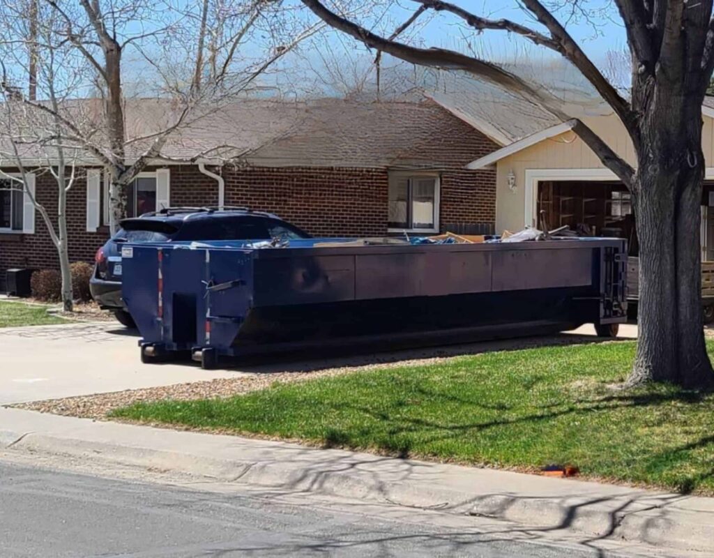 Large blue dumpster in a residential driveway beside a brick house, ready for waste disposal or renovation cleanup.