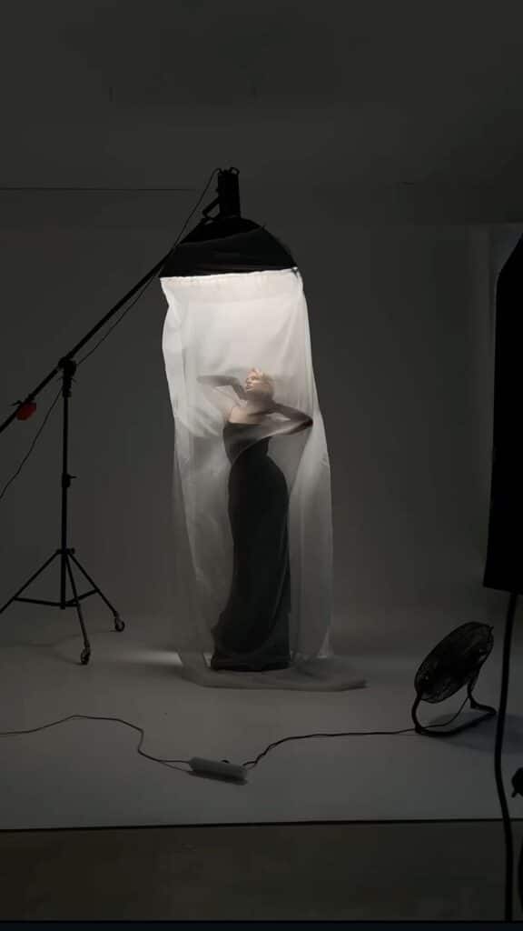 Person posing dramatically under soft light and sheer fabric, highlighting fashion photography in a studio setup.
