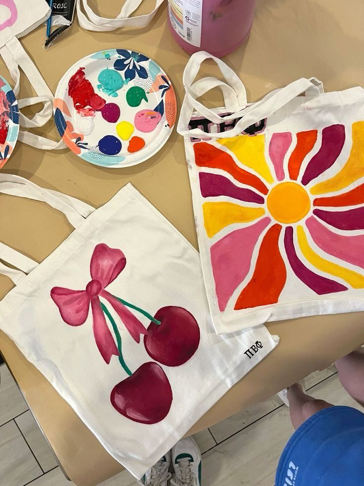 Hand-painted tote bags with cherry and sunburst designs next to colorful paint palette on a table.