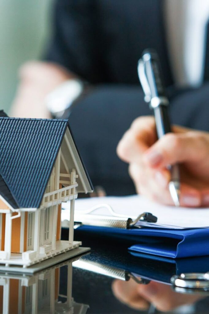 40f07d4f-9e72-4b59-a8d9-65987ba7157b Person signing real estate document with house model on table. | Sky Rye Design Person signing real estate document with house model on table.