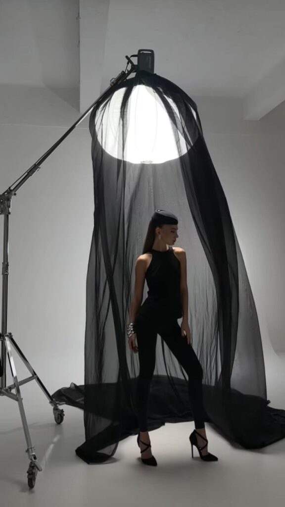 Fashion model under dramatic lighting with a sheer black drape and a hat, wearing a stylish black outfit and heels.