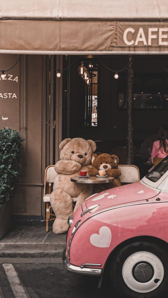 Teddy bears at a café table with coffee, beside a pink car with heart designs, creating a whimsical street scene.