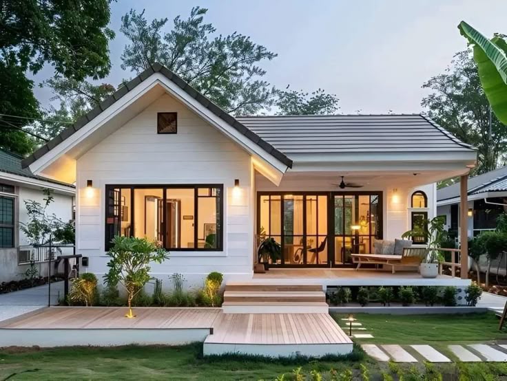 Modern white house with a spacious porch, large windows, and surrounding greenery at dusk.