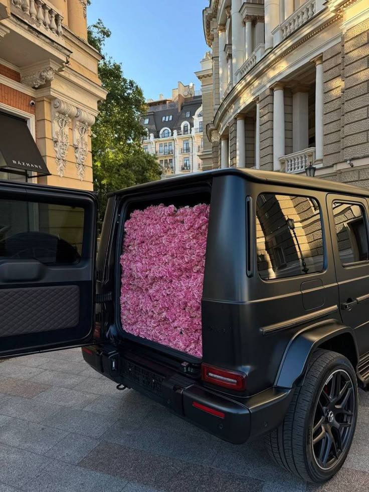 Black SUV trunk overflowing with pink roses, parked in front of elegant buildings on a sunny day.