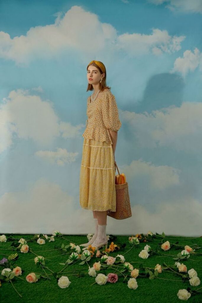 Woman in yellow floral dress with basket, standing on grass with flowers and cloudy sky background.