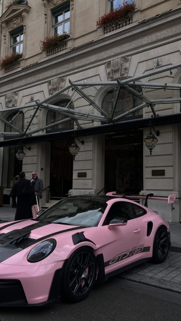 Pink sports car parked outside an elegant building with flowers in window boxes above.