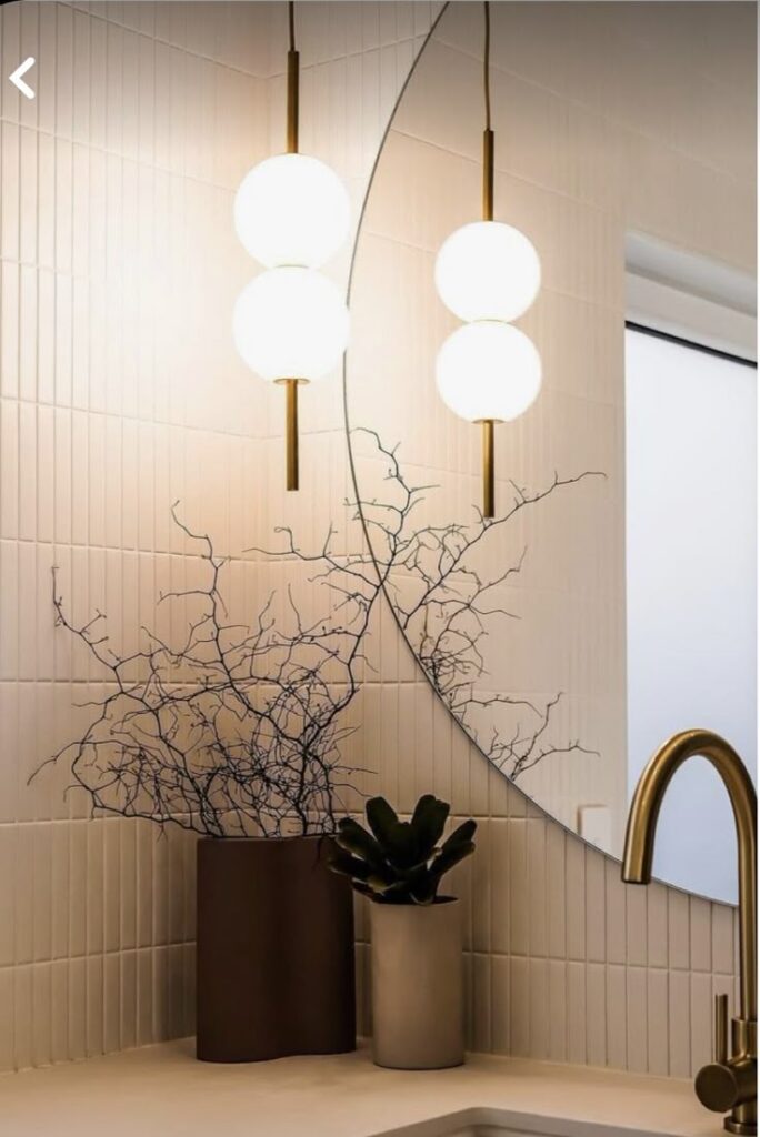 Modern bathroom with round mirror, gold faucet, pendant lights, and elegant plants in vases on a tiled counter.