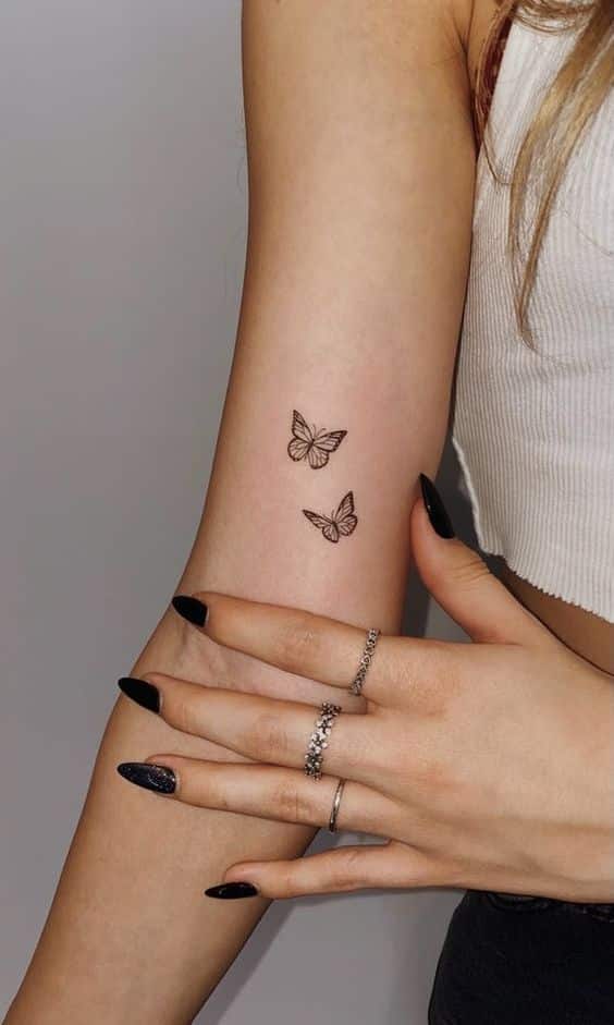 Two small butterfly tattoos on a person's arm, with black nails and rings, wearing a white tank top.