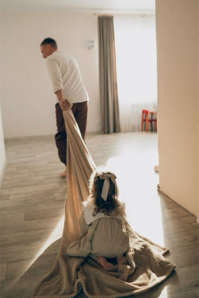 Father playfully pulls daughter on blanket through sunlit room.