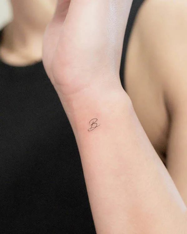 Minimalist B wrist tattoo on a person wearing a black top.